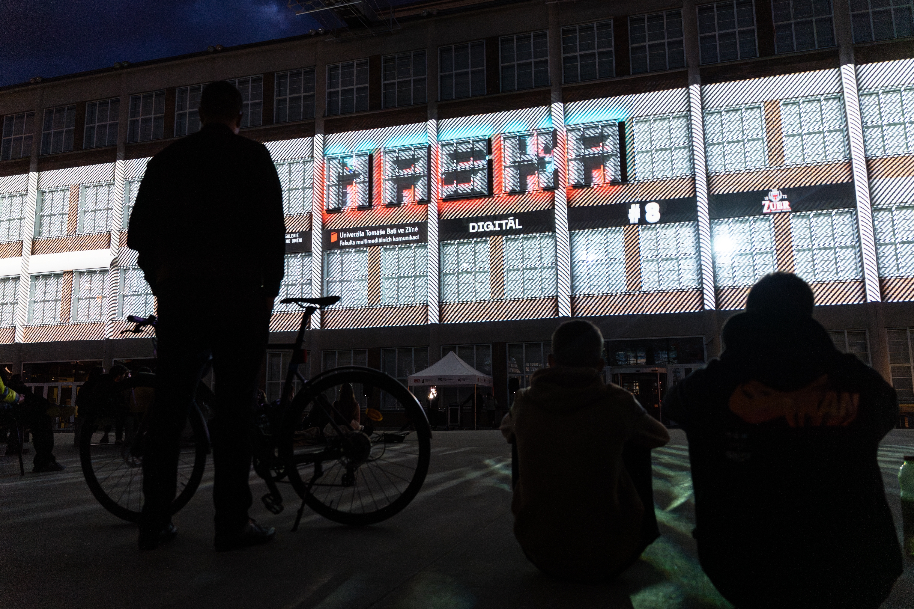 foto z videomappingu MASKA, v popředí tři osoby dívající se na videomapping na zdi budovy